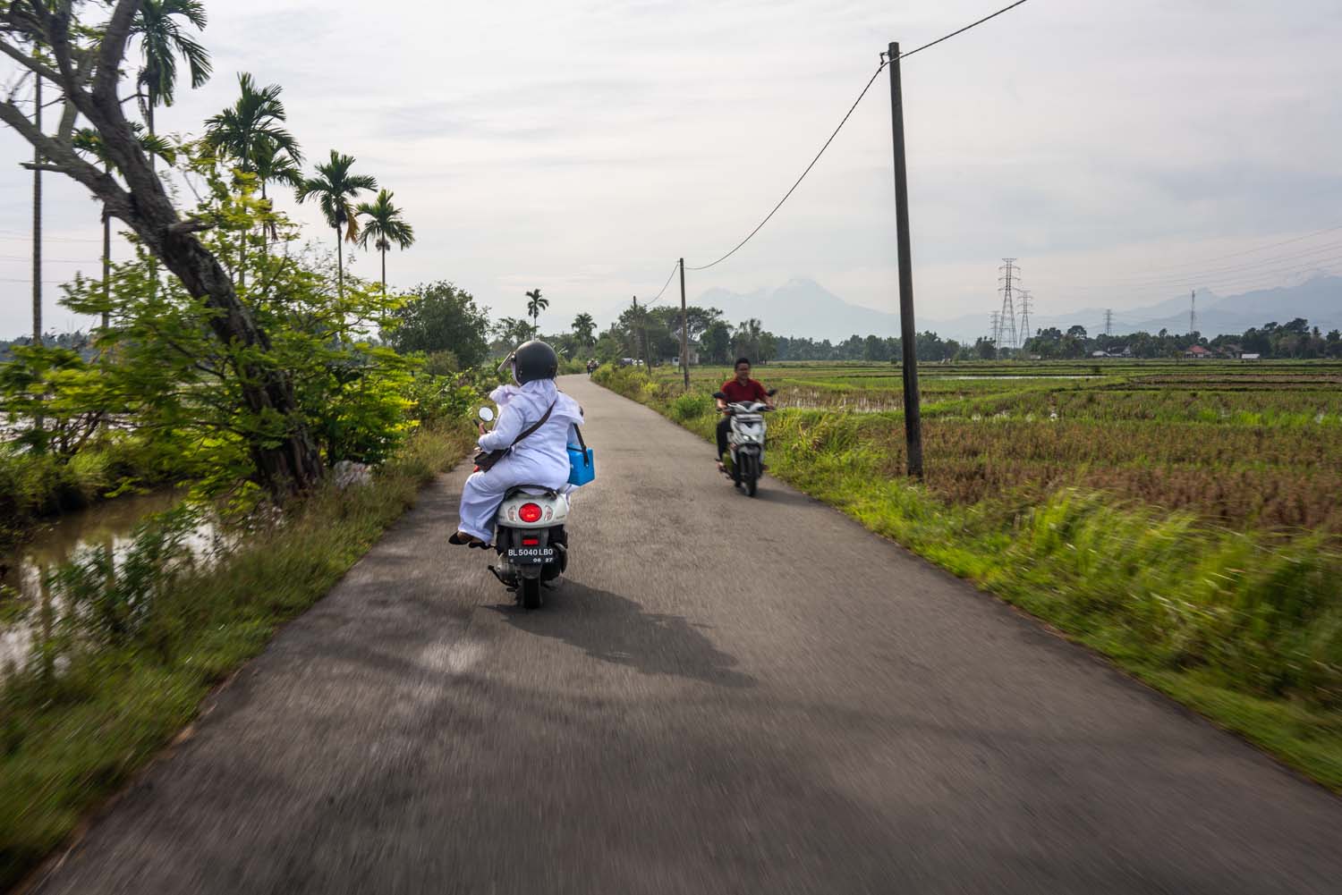 Two midwives riding a motorbike on an asphalt road in Aceh Besar District, Aceh Province. On their right and left are green paddy fields.